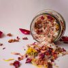 A glass jar of aromatic bath salts mixed with dried flowers and herbs spilled over a white background.